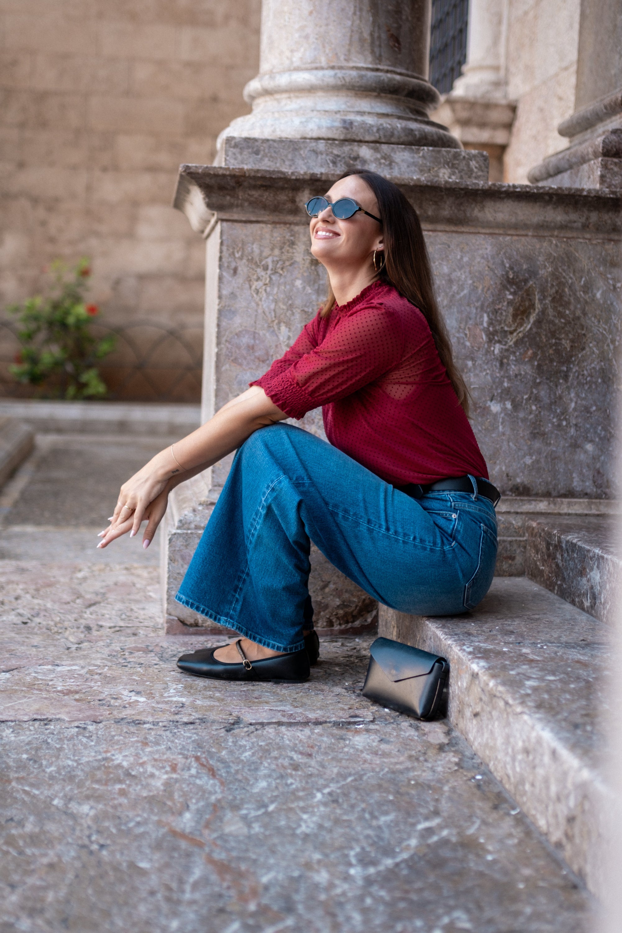 Woman sitting on stone steps wearing sunglasses and a red shirt with blue jeans.
