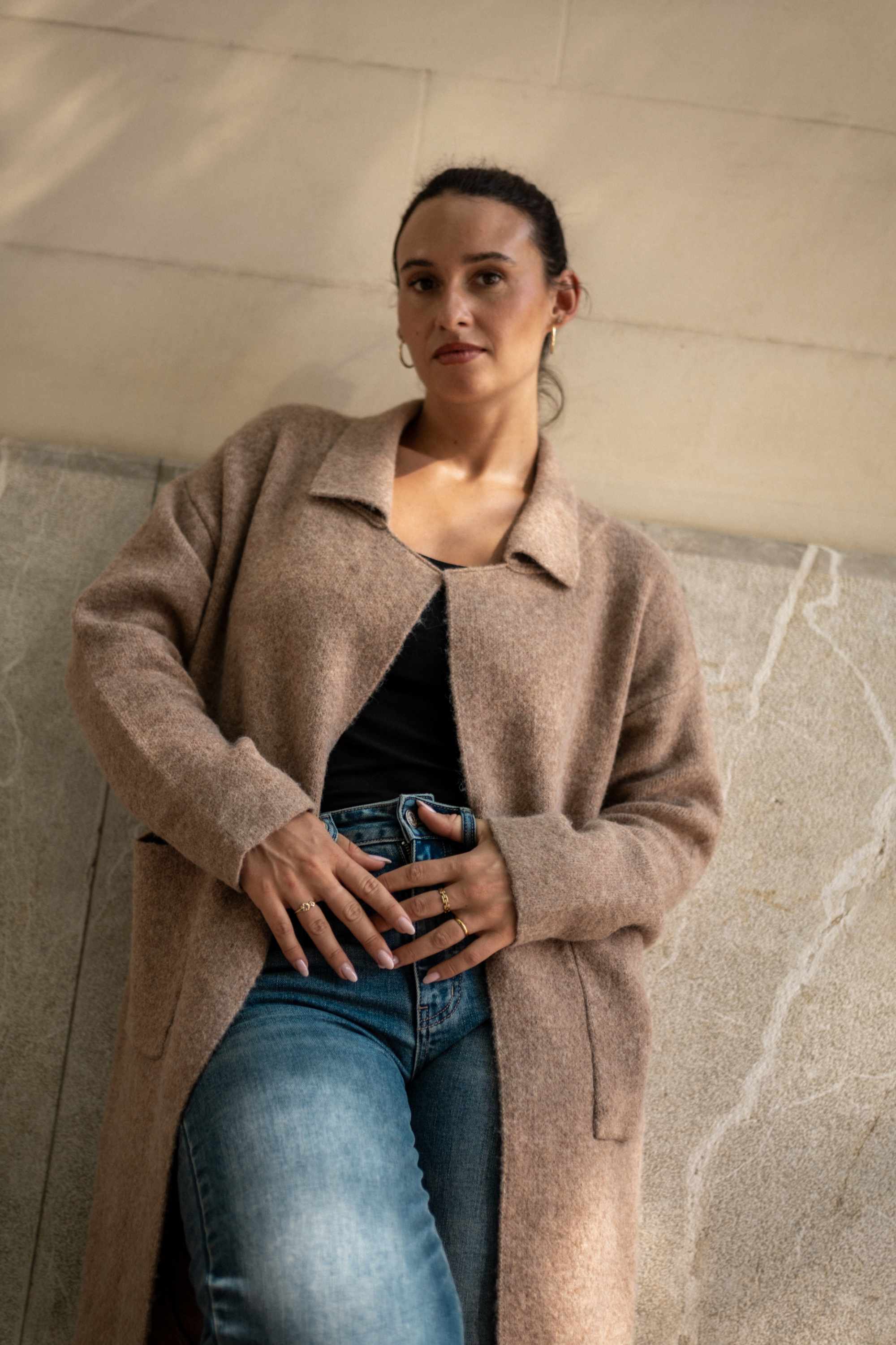 Woman wearing a long beige coat against a marble wall.