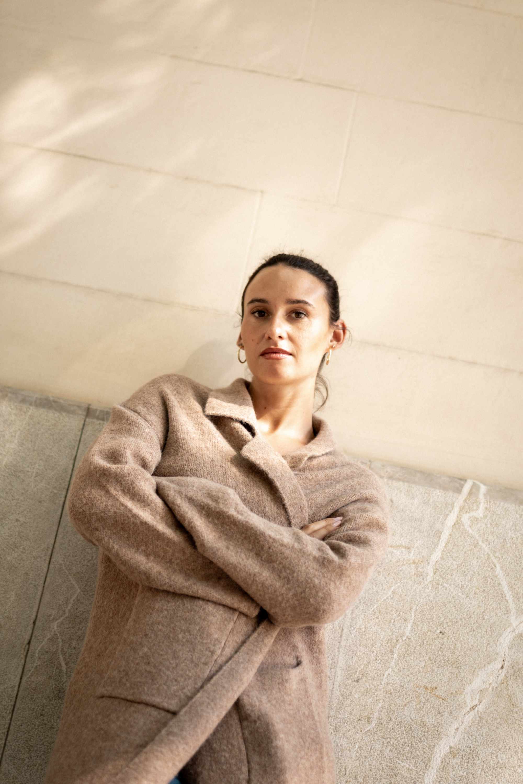 Woman wearing a beige coat standing against a neutral wall.