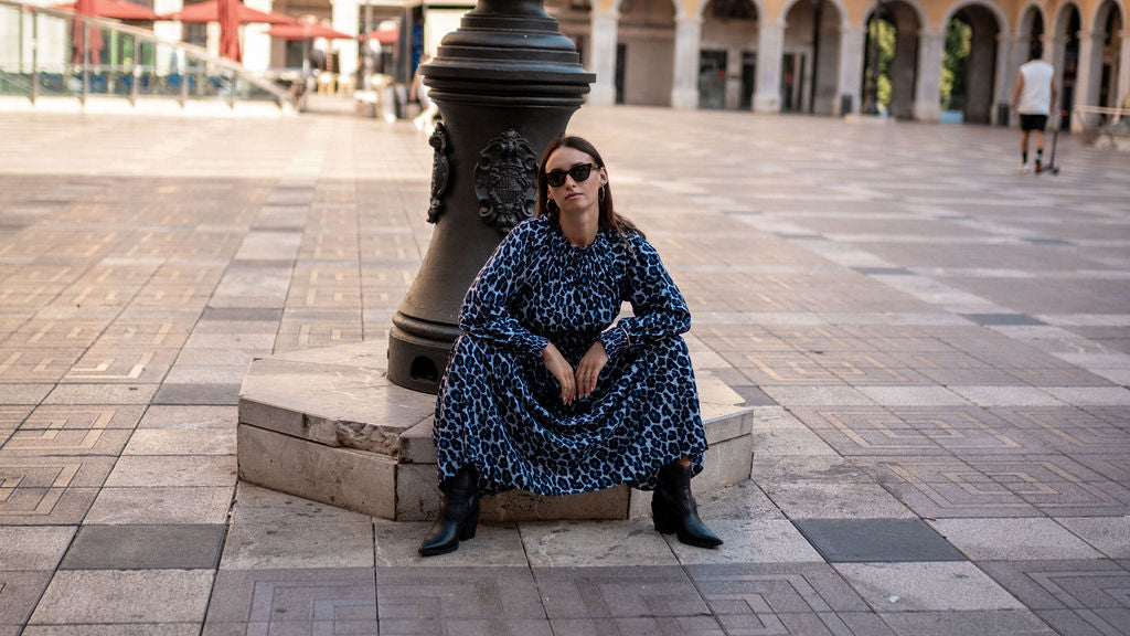 Woman in a blue floral dress sitting on a stone bench in an outdoor square.