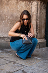 Woman in black sleeveless top and blue jeans holding a black clutch against a stone wall.