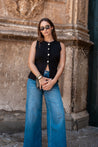 Woman wearing a black sleeveless top and blue jeans standing in front of an ornate stone wall.