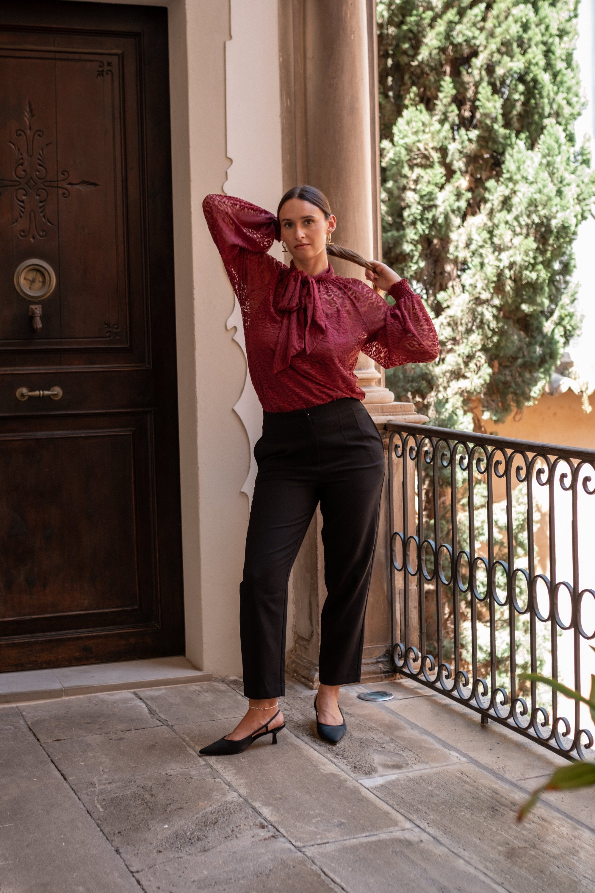 Woman in a burgundy lace blouse and black pants standing on a stone patio with a wooden door and greenery in the background.