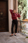 Woman in a burgundy lace blouse and black pants standing on a stone patio with a wooden door and greenery in the background.
