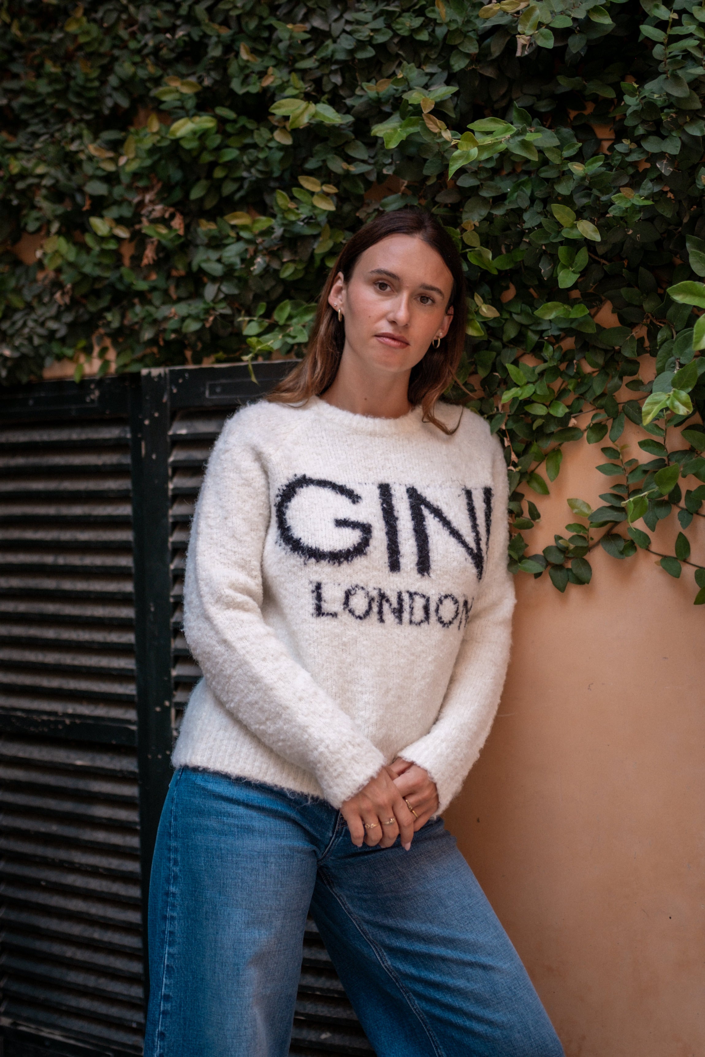 Person wearing a white sweater with 'GINI LONDON' text against a green leafy background