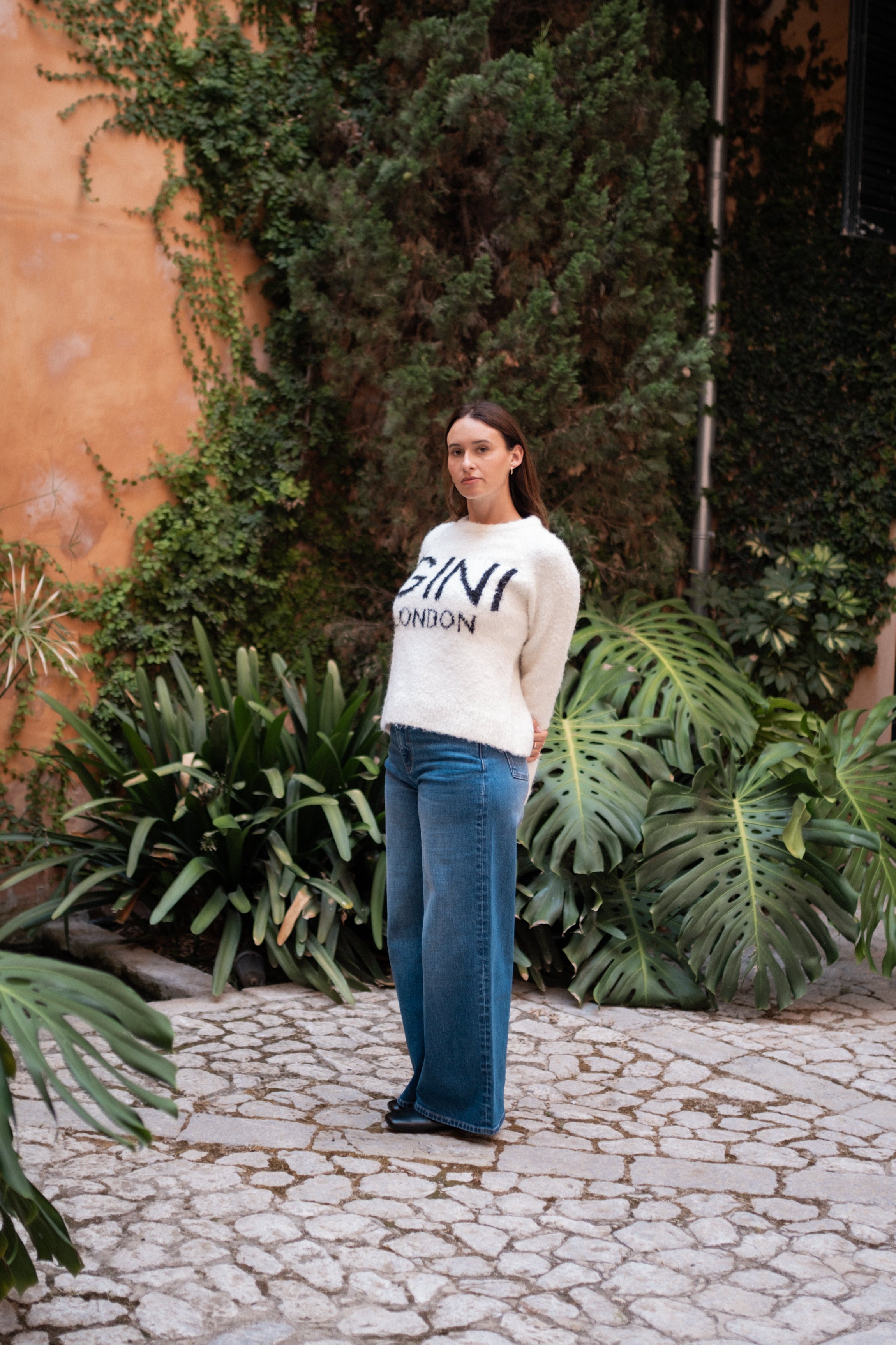 Woman wearing a white sweatshirt with text standing on a stone path with greenery.
