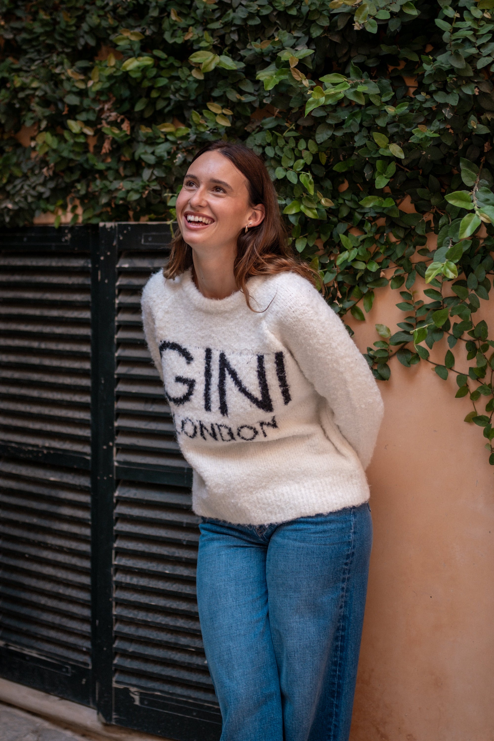 Woman wearing a white sweater with 'GINI LONDON' text, standing against a wall with greenery.
