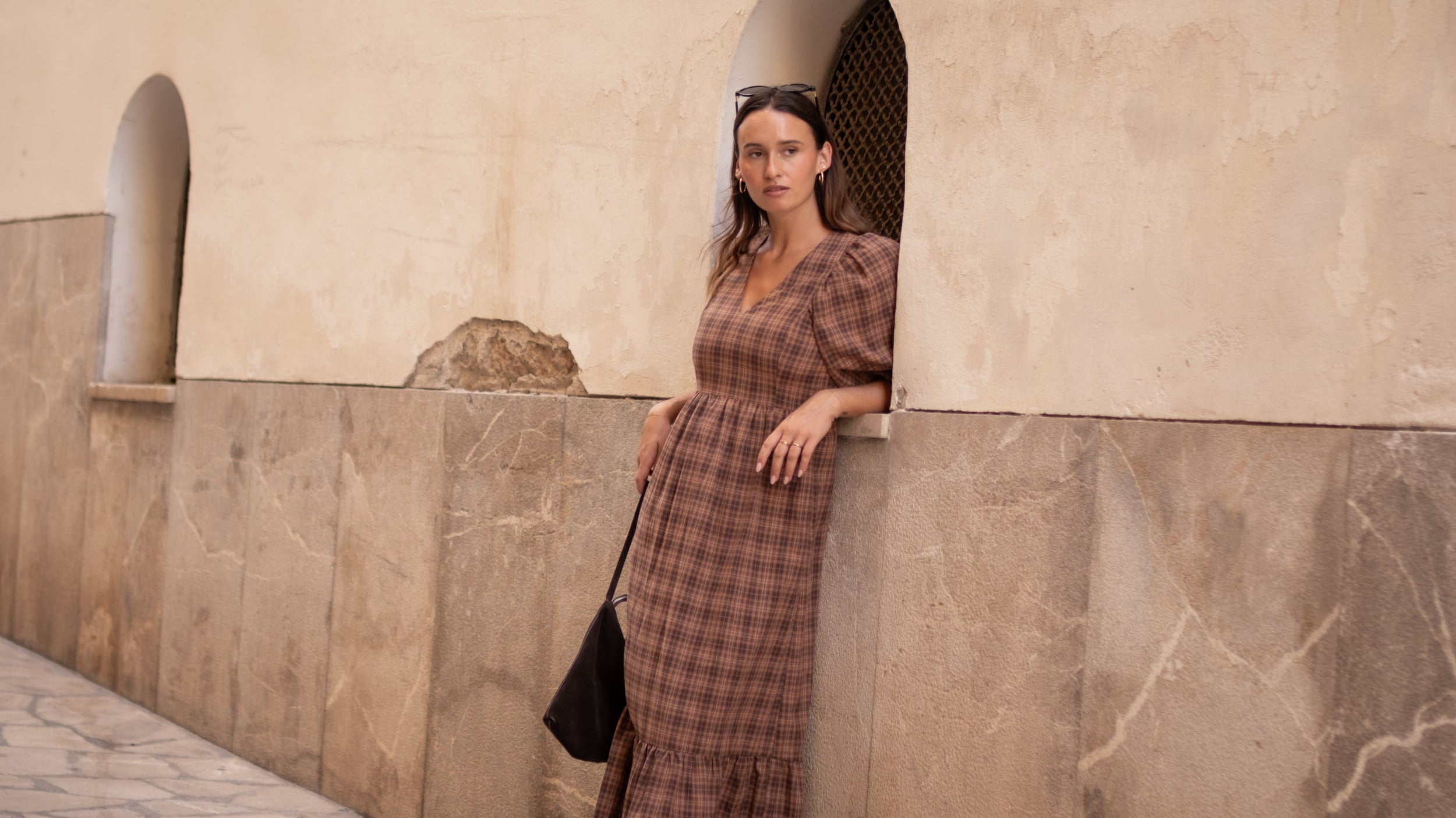 Woman in a plaid dress standing against a stone wall with arched windows.